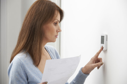 A woman adjusting the thermostat in a home while holding an energy bill.