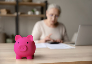 A woman sitting in front of a laptop with a calculator. There is a piggy bank in the foreground.