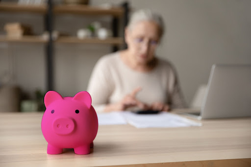 A woman sitting in front of a laptop with a calculator. There is a piggy bank in the foreground.