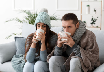 A man and woman looking cold as they sit on a couch and drink out of mugs.