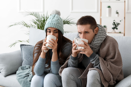 A man and woman looking cold as they sit on a couch and drink out of mugs.