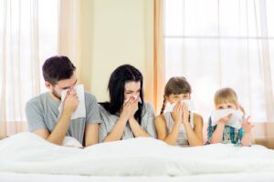 A family of four sitting up in a bed and blowing their noses due to poor IAQ.