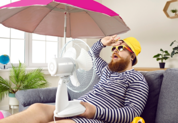 Man sitting on the couch with a fan and an umbrella, touching his forehead in exasperation because AC is blowing warm air.