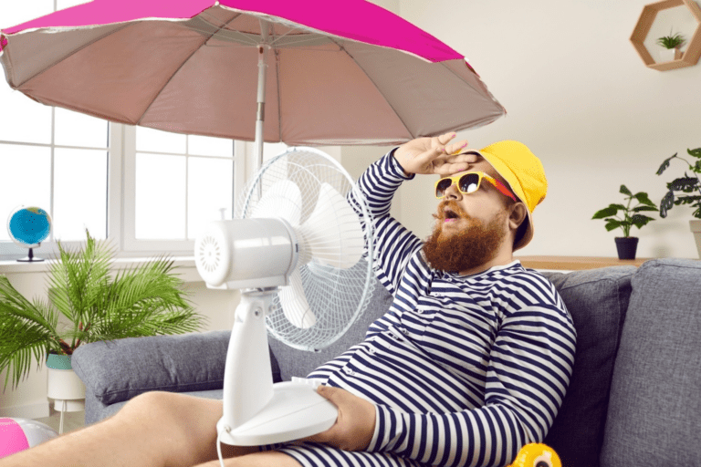 Man sitting on the couch with a fan and an umbrella, touching his forehead in exasperation because AC is blowing warm air.