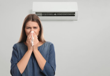 A woman blowing her nose as she stands in front of a wall-mounted AC unit.