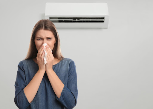 A woman blowing her nose as she stands in front of a wall-mounted AC unit.