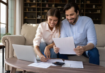 A man and woman sitting on a couch in a home and looking over papers with a calculator and pen.