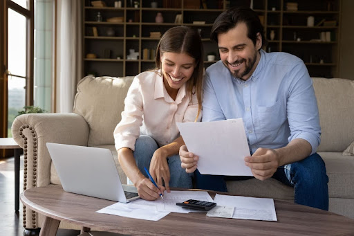 A man and woman sitting on a couch in a home and looking over papers with a calculator and pen.