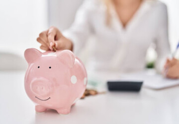 A woman sitting at a desk with a pen, paper, and calculator; she's putting a coin into a piggy bank.