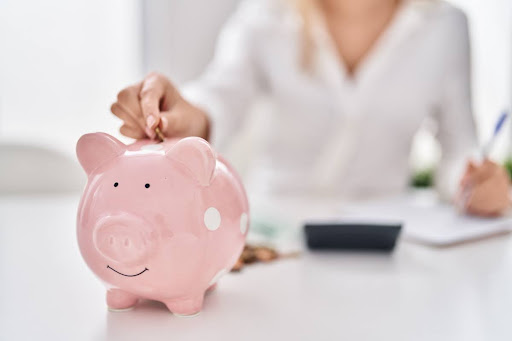 A woman sitting at a desk with a pen, paper, and calculator; she's putting a coin into a piggy bank.