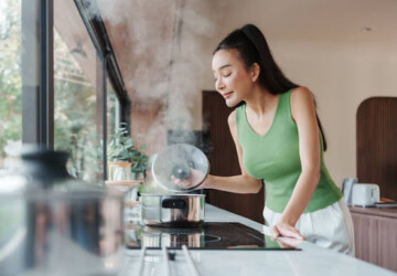 A woman standing in a kitchen, boiling water, and smelling the steam rising from a pot.