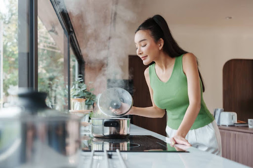 A woman standing in a kitchen, boiling water, and smelling the steam rising from a pot.